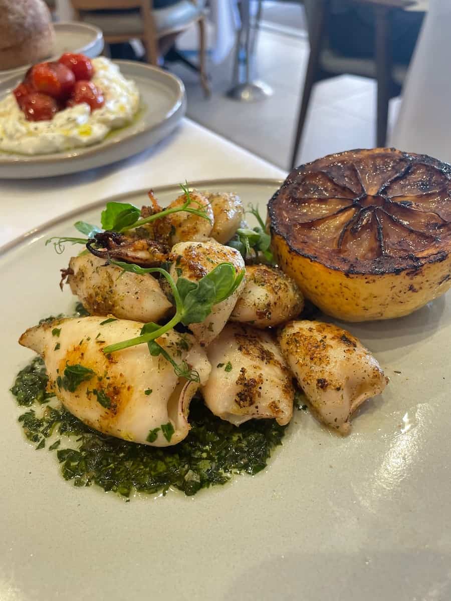 Grilled octopus with a burnt lemon and a plate of whipped feta and tomatoes in the background